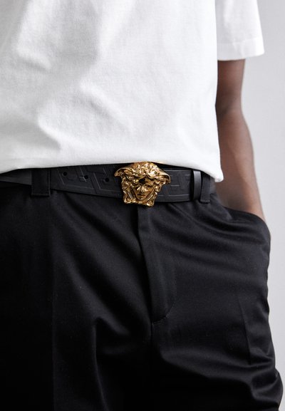 Cinturón negro con una prominente hebilla con cabeza de león dorada, superficie texturizada con patrones geométricos, usado sobre pantalones oscuros y una camisa blanca.