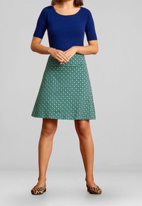 Blaues kurzärmeliges Oberteil kombiniert mit einem grünen gemusterten A-Linie-Rock mit geometrischen Designs. Dazu werden Ballerinas im Leopardenmuster getragen.