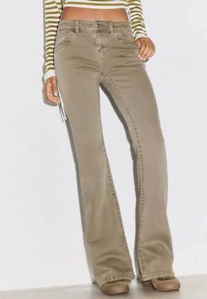 Femme portant un pantalon évasé beige, un crop top à manches longues rayé vert et blanc, et des chaussures beige, debout sur un fond blanc.