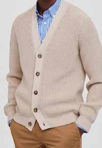 Beżowy sweter dzianinowy z dekoltem w kształcie litery V, z brązowymi guzikami, prążkowanymi mankietami i dołem, noszony na niebieskiej i białej pasiastej koszuli.