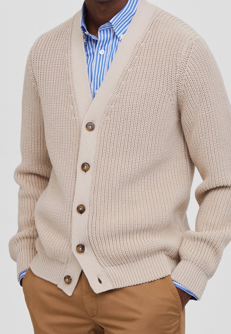 Beżowy sweter dzianinowy z dekoltem w kształcie litery V, z brązowymi guzikami, prążkowanymi mankietami i dołem, noszony na niebieskiej i białej pasiastej koszuli.