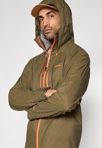 Αδιάβροχο τζάκετ σε ελιά πράσινο με πορτοκαλί φερμουάρ και λεπτομέρειες, ψηλό γιακά με κουκούλα και ρυθμιζόμενα μανσέτα. Ελαφρύ, με απαλή υφή.