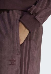 Adidas sweatpants i en mörk plommonfärg, med en mjuk textur, ribbade muddar, sidofickor och tre vertikala ränder längs sidorna.