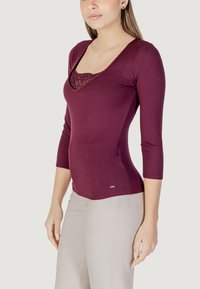 Top a maniche lunghe bordeaux con texture a coste e dettaglio in pizzo sullo scollo, design aderente, abbinato a pantaloni di colore chiaro.