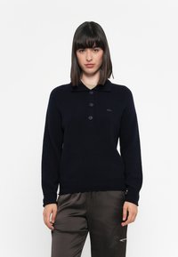 Maglione lavorato a maglia blu navy con colletto, chiusura a tre bottoni e orlo a coste aderente. Logo Lacoste sul lato sinistro del petto.