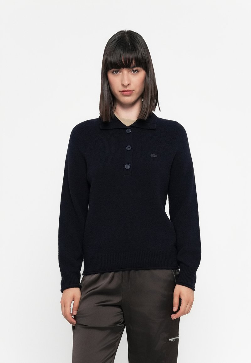 Maglione lavorato a maglia blu navy con colletto, chiusura a tre bottoni e orlo a coste aderente. Logo Lacoste sul lato sinistro del petto.