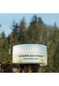 Bocal en plastique blanc avec un couvercle transparent, illustré d'oiseaux jaunes. Le texte à l'avant indique "SUSANNE KAUFMANN Beurre corporel pour les sens."