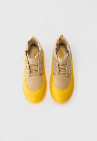 Bottes imperméables beiges et jaunes avec lacets, doublure en fourrure, et embouts en caoutchouc texturé jaune, vues d'en haut sur un fond blanc.