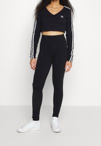 Svart kortärmad långärmad topp med knappstängning och vita ränder på ärmarna, kombinerad med högmidjade svarta leggings och vita sneakers.