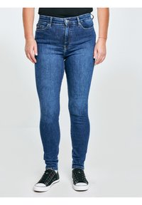 Inimene, kes kannab tumesiniseid kitsaid teksapükse ja musti tossusid, seisab ühevärvilise heleda tausta ees.