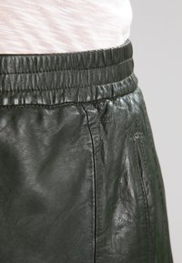 Tmavě zelená kožená sukně s hladkým povrchem, elastickým pasem a jemným leskem. Obsahuje boční kapsy a precizní prošívání.