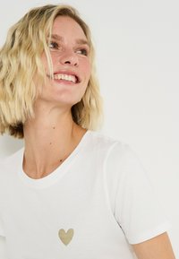 Camiseta blanca de manga corta hecha de tela suave, con un pequeño diseño de corazón dorado en el área del pecho izquierdo y un escote redondo.