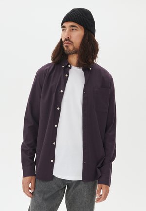 Mand med langt hår og skæg iført sort beanie, mørk lilla skjorte med knapper over hvid t-shirt og grå jeans, der står foran en hvid baggrund.