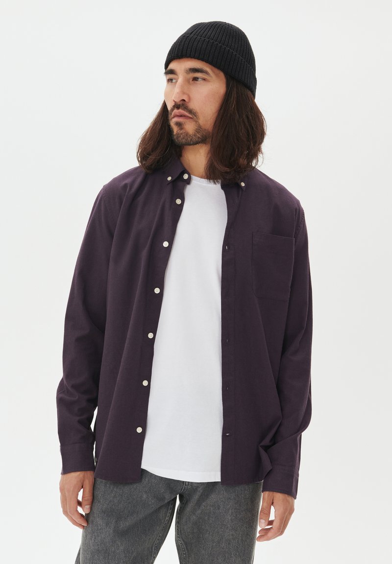 Man med långt hår och skägg som bär en svart mössa, en mörk lila skjorta över en vit t-shirt och grå jeans, stående mot en vit bakgrund.