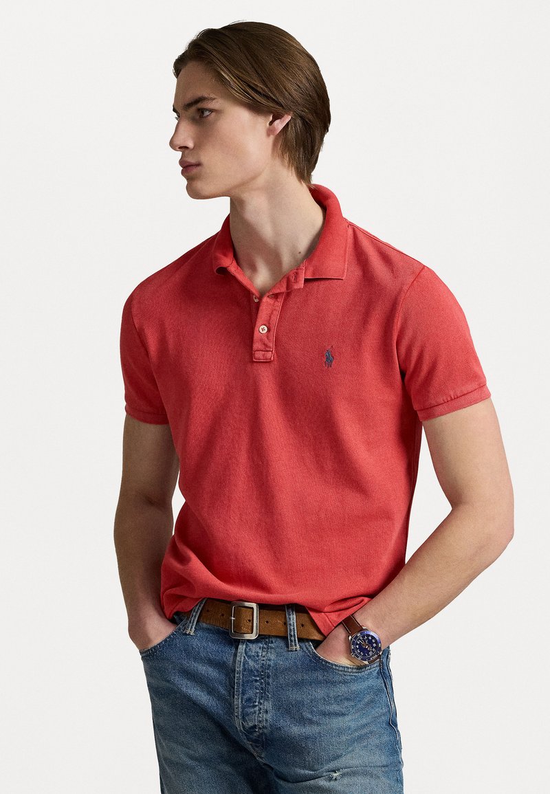 Rød bomullspolo med klassisk krage og korte ermer, med en liten brodert logo. Brukt med blå jeans og et brunt belte.