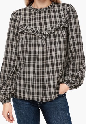 Kvinde iført sort og grå ternet langærmet bluse med flæsedetalje over brystet og ærmer med manchetter, kombineret med blå jeans.