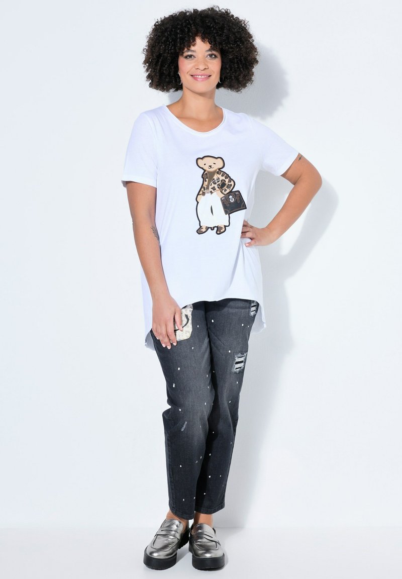 Frau mit lockigem Haar trägt weißes T-Shirt mit Bär-Grafik, zerrissene schwarze Jeans mit weißen Punkten und metallisch glänzende Slip-On-Schuhe, steht vor einer weißen Wand.