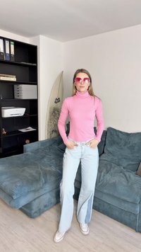 Rosa turtle-neck genser, lyseblå jeans med sidesplitter, og rosa solbriller. I bakgrunnen er det en blå sofa og hyller med diverse gjenstander.