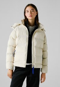 Puffermantel in hellem Creme, mit hohem Kragen, Reißverschluss und blauem Reißverschluss-Zipper. Strukturierte Steppdesign mit Seitentaschen.