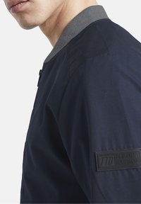 Námořnická bombardovací bunda s šedým žebrovaným límcem, se strukturovanou tkaninou a černou značkovou nášivkou na levém rukávu.