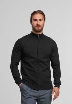Schwarzer Reißverschluss-Pullover aus strukturiertem Stoff, mit hohem Kragen und gerippten Bündchen, getragen über einer hellen, gemusterten Hose.