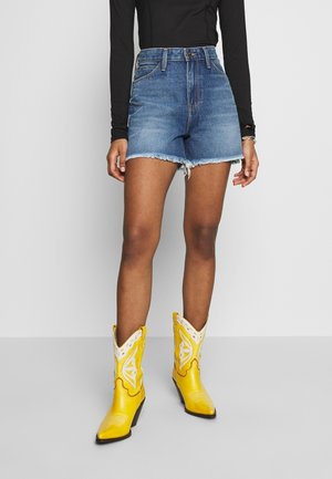 Shorts en denim taille haute avec un ourlet effiloché de couleur bleu moyen. Associés à des bottes de cowboy jaune vif ornées de détails découpés blancs complexes.