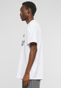 T-shirt en coton blanc à manches courtes, arborant un texte noir sur le devant. Le t-shirt a une coupe décontractée et est associé à un pantalon gris.