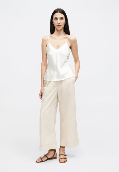 Žena s dlouhými tmavými vlasy, oblečená v bílém tílku bez rukávů, béžových širokých pruhovaných kalhotách a hnědých plochých sandálech, stojící s jednou rukou v kapse.