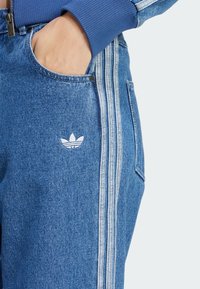 Denim joggers in light blue with three white side stripes. Features a front pocket and an embroidered white logo near the pocket.