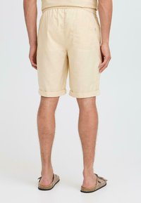 Lys beige shorts laget av mykt stoff, med oppbrettet kant, elastisk midje og baklomme. Bært med brune sandaler.