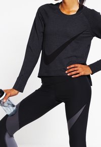 Maglia sportiva nera a maniche lunghe con texture a coste e un grande logo Nike nero. Abbinata a leggings neri con accenti grigi e una bottiglia d'acqua.