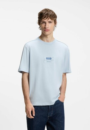 Junger Mann in hellblauem HUGO T-Shirt mit Logo-Patch, Hände in den Taschen, trägt dunkelblaue Jeans, steht vor weißem Hintergrund.