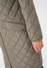 Îmbrăcăminte exterioară matlasată de culoare gri cu un model texturat, cu un fermoar lateral și un buzunar cu buton de fixare. Materialul pare moale și ușor.