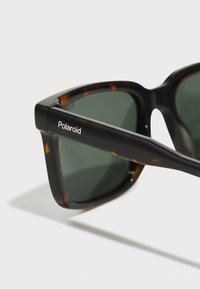 Occhiali da sole rettangolari neri e tartarugati con lenti verdi, con logo "Polaroid" bianco sulla tempia. Montatura in plastica liscia.