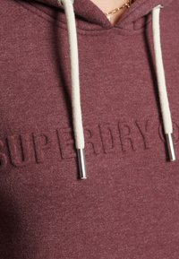 Bordeaux hoodie met een getextureerd "SUPERDRY" logo, voorzien van crème kleurige koordjes met metalen punten. Zachte, fleece-achtige stof voor comfort.