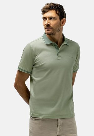 Man met kort krullend haar en baard, draagt een lichtgroen poloshirt en beige broek, staat met handen achter de rug, kijkt opzij.