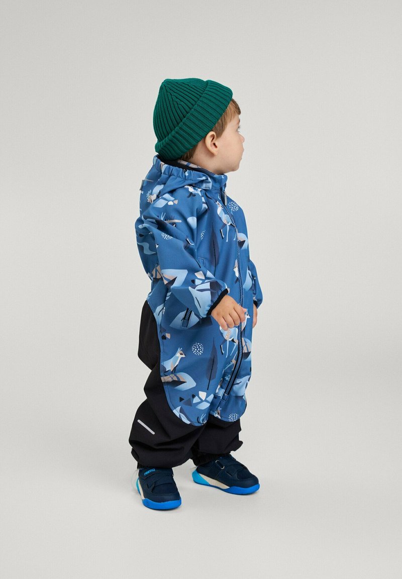 Blauwe waterdichte jas met dierenpatronen, gecombineerd met zwarte broek. Een groene gebreide beanie maakt de outfit compleet. Blauwe en zwarte schoenen worden gedragen.