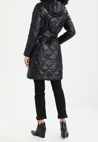 Svart, quiltad pufferjacka med huva och fuskpälsdekoration, åtdragen i midjan med ett bälte, kompletterad med tajta svarta byxor och ankelstövlar.