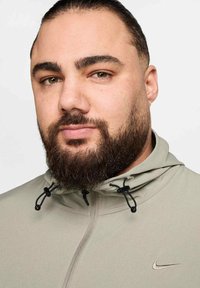 Nike-hoodie in lichtgrijs met een ritssluiting, verstelbare trekkoorden en een subtiel logo op de borst. Zachte, gladde stoftextuur.