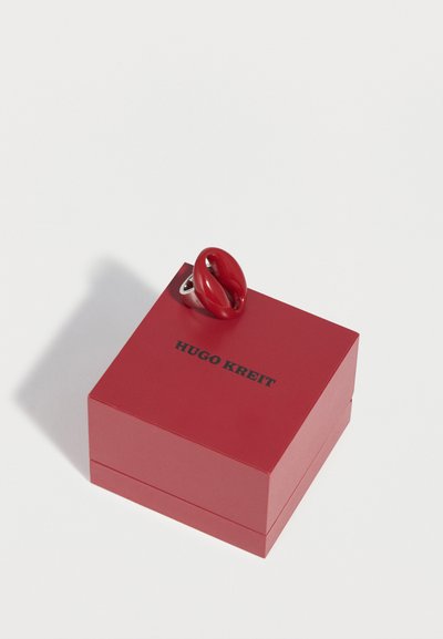 Bague en argent avec motif de lèvres rouges reposant sur une boîte à bijoux rouge fermée Hugo Kreit sur fond blanc.