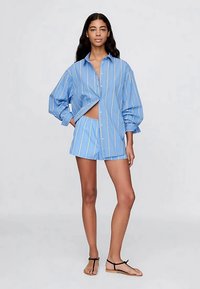 Vrouw met lang, donker haar, die een losse blauw gestreepte blouse en bijpassende shorts draagt, met zwarte sandalen op een effen achtergrond.