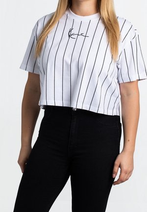 Kobieta ubrana w biały krótki T-shirt z pionowymi czarnymi paskami oraz czarne spodnie z wysokim stanem, stojąca na jednolitym tle.