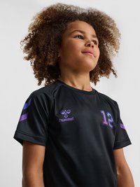 Young person with curly hair wearing a black sports jersey with number 12 and purple striped details on sleeves.