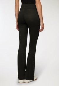 Kobieta w czarnych dopasowanych spodniach w stylu dzwonów i białych trampkach, stojąca tyłem na jednolitym jasnym tle.