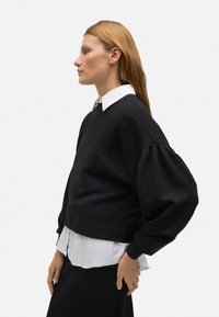 Sudadera corta negra con mangas voluminosas, superpuesta a una camisa blanca con cuello, con un diseño sencillo y una textura suave.