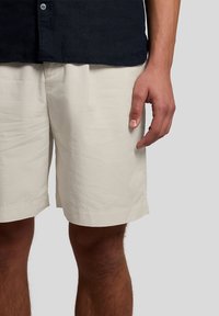 Άνδρας που φοράει μπεζ σορτς μέχρι το γόνατο και σκούρο μπλε πουκάμισο, με το δεξί χέρι χαλαρό στο πλάι του, μπροστά σε απλό φόντο.