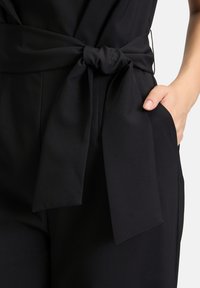 Fekete overál, megkötős derék övvel, lapos elülső zsebekkel és sima, texturált anyaggal. Közeli nézet a derék területéről.