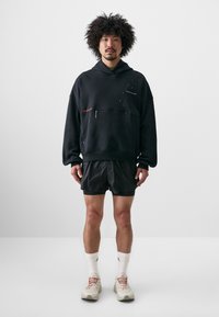Sort hætte sweatshirt med lynlås-lomme, parret med sorte shorts. Har refleksdetaljer og strukturerede sneakers. Hvide crew-sokker synlige.