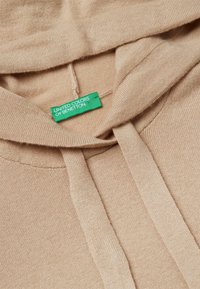 Hoodie beige avec une texture douce, doté d'une poche kangourou à l'avant et de cordons de serrage ; l'étiquette indique "UNITED COLORS OF BENETTON" en vert.