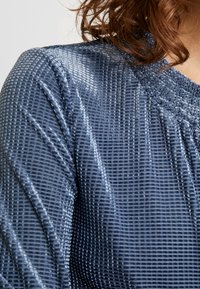 Blaues strukturiertes Gewebe mit einem Rautenmuster, das einen subtilen Glanz aufweist. Die gerippte Ausschnitt- und Detailverarbeitung verstärkt das Design.